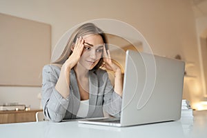 Tired woman sitting at desk with laptop, holding head, resting on hand.