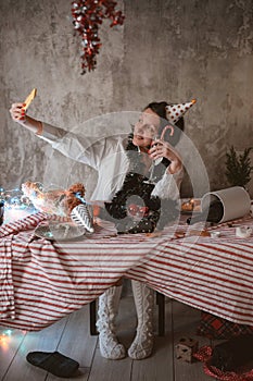 Tired woman taking selfie on mobile phone while sitting at unmade decorated New Years festive table