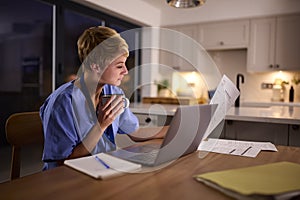 Tired Woman Wearing Medical Scrubs Working Or Studying On Laptop At Home At Night