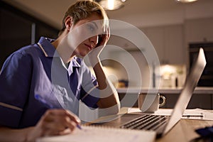 Tired Woman Wearing Medical Scrubs Working Or Studying On Laptop At Home At Night