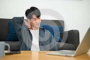 Tired young Asian male freelancer sits in the living room using laptop, touching his neck