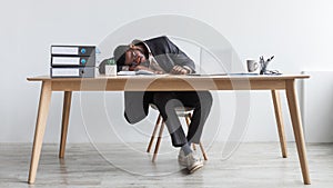 Tired young black businessman sleeping on desk in front of laptop, overworking, feeling exhausted at office, full length