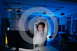 Tired young businessman stretching in front of computer sitting on desk at office workplace at late night in dark room