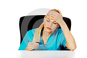 Tired young female doctor or nurse sitting behind the desk and holding termomether