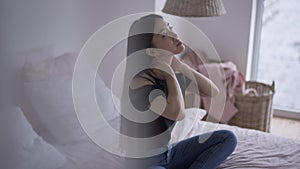 Tired young female freelancer stretching neck sitting on bed indoors. Side view wide shot of Asian woman suffering pain