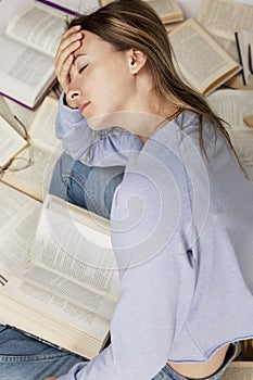 A tired young girl sits among a large pile of opened books. Training and education. Vertical