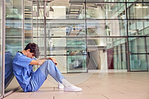 Tired Young Male Doctor Wearing Scrubs Sitting Against Wall In Modern Hospital Building