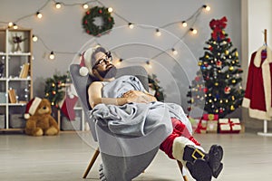 Tired young man dressed as Santa Claus sleeping in an armchair under warm blanket