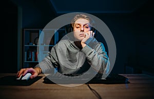 Tired young man in headset playing video games at home on computer in room with blue light and looking at camera with sleepy face