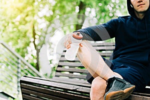 Tired Young man runner in hoodie sitting on bench and relaxing after sport training. Holding water bottle while doing