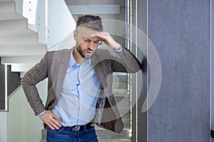 Tired young man standing by the wall in the office and holding his head, feeling bad, worried