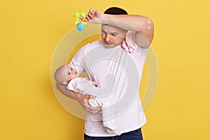 Tired young man in white casual t shirt holding newborn daughter in hands, looks exhausted and sleepy, keeps hand with beanbag