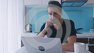 Tired young man working on a laptop at home in the kitchen. A man suffers and has a headache.