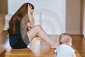 Tired young mother sits in the bedroom on the floor, a half-year-old baby crawls nearby