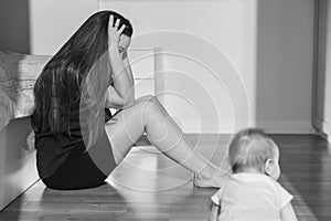 Tired young mother sits in the bedroom on the floor, a half-year-old baby crawls nearby