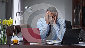 Tired young overworking man messaging online on laptop and tablet holding head in hands looking at camera with exhausted