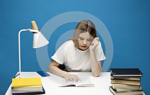 Tired young student woman in white casual clothes sitting at the table reading the book in library of university or