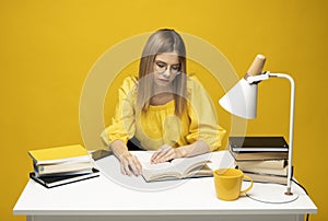 Tired young student woman in yellow casual clothes sitting at the table reading the book in library of university or