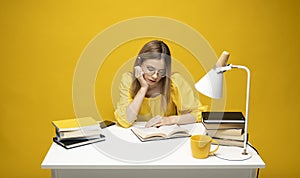 Tired young student woman in yellow casual clothes sitting at the table reading the book in library of university or