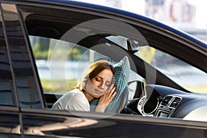 Tired young woman asleep on pillow on steering wheel, resting after long hours driving a car. Fatigue