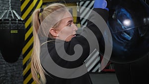 A tired young woman in blue boxing gloves hits a punching bag in the gym.