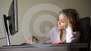 Tired young woman with curly hair and white shirt is working at the office using her computer, routine work, freelance