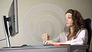 Tired young woman with curly hair and white shirt is working at the office using her computer, routine work, freelance