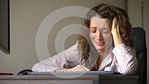 Tired young woman with curly hair and white shirt is working at the office using her laptop, routine work, freelance