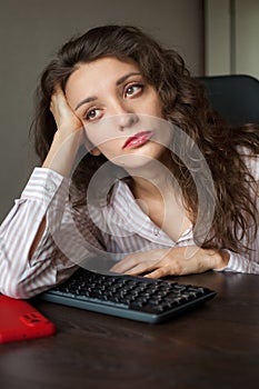 Tired young woman with curly hair and white shirt is working at the office using her laptop, routine work, freelance