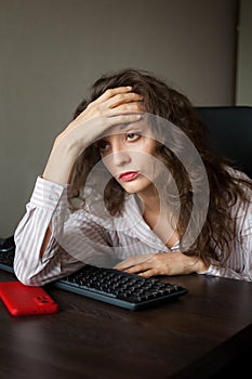 Tired young woman with curly hair and white shirt is working at the office using her laptop, routine work, freelance