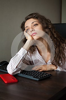 Tired young woman with curly hair and white shirt is working at the office using her laptop, routine work, freelance