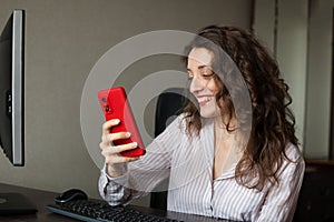 Tired young woman with curly hair and white shirt is working at the office and using her red smartphone, routine work