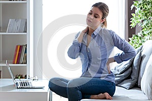 Tired young woman with shoulder and back pain sitting on the couch at home.