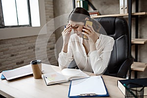 Tired young woman sit at table in room. She talk on phone and hol hand on forehead. Tired and exhaustion.