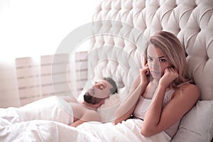 Tired young woman sitting in bed with snoring husband