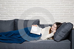 Tired young woman taking a nap at home lying on a sofa with a book lying across her chest and her eyes closed