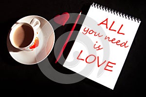 Title ALL you need is love on the notebook blank with cup of coffee, red heart on the black background. Top view with copy space.