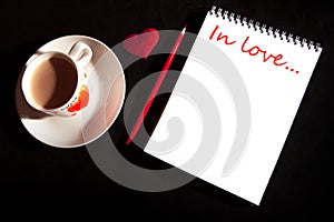 Title In love... on the notebook blank with cup of coffee, red heart on the black background. Top view with copy space. Love and