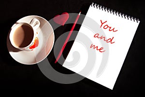 Title You and me on the notebook blank with cup of coffee, red heart on the black background. Top view with copy space. Love and