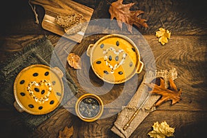 To bowls with pumpkin soup with cream and seeds on a rustic metal and wooden background, decorated with autumn leaves