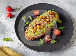 Toast with avocado, roasted chickpeas, cherry tomatoes, herbs on a dark plate.