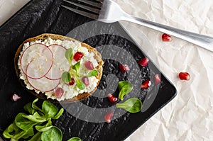 Toast with cottage cheese and smashed avocado, radish, corn salad plant and pomegranate seeds on black plate.