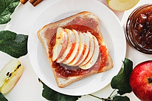 Toast with a crushed apple jam, slices of fresh apples, cinnamon on a plate on a white table. Apples and apple jam in jar. The con