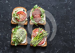 Toast sandwiches with avocado, salami, asparagus, tomatoes and soft cheese on dark background, top view. Tasty breakfast, snack or