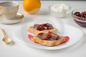 Toasted French bread, plum jam, cottage cheese and orange on a white wooden table.