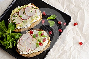 Toasts with cottage cheese and smashed avocado, radish, corn salad plant and pomegranate seeds on black plate.