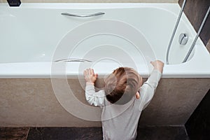 Toddler baby boy climbs into the white tub standing at the edge. Child plays in the bathroom, kid and bath