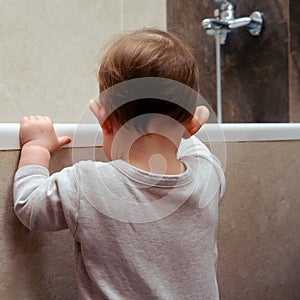 Toddler baby boy climbs into the white tub standing at the edge. Child plays in the bathroom, kid and bath