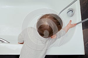 Toddler baby boy climbs into the white tub waddling over the edge. Child plays in the bathroom, kid and bath