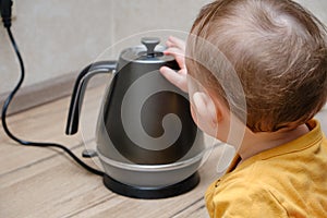 Toddler baby boy holds a hot kettle with boiling water. Child safety issues in the home room, little kid